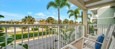 A balcony with white railings, a white chair with a striped pillow, and a view of a street with palm trees and houses.