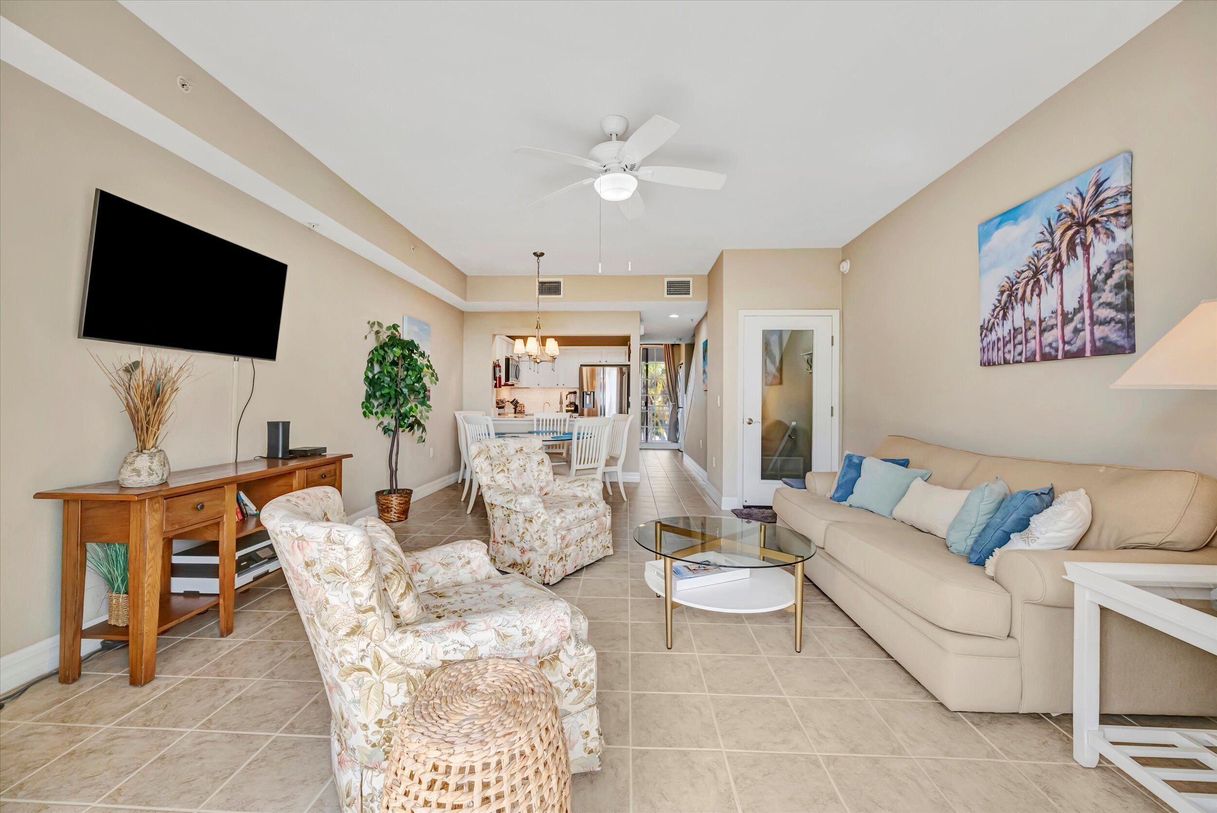 A living room with a flat-screen TV, a ceiling fan, a dining area with a chandelier, and a painting of palm trees.