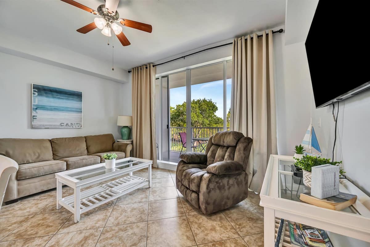 A living room with a sofa, a recliner, a coffee table, a TV, and a balcony with curtains.