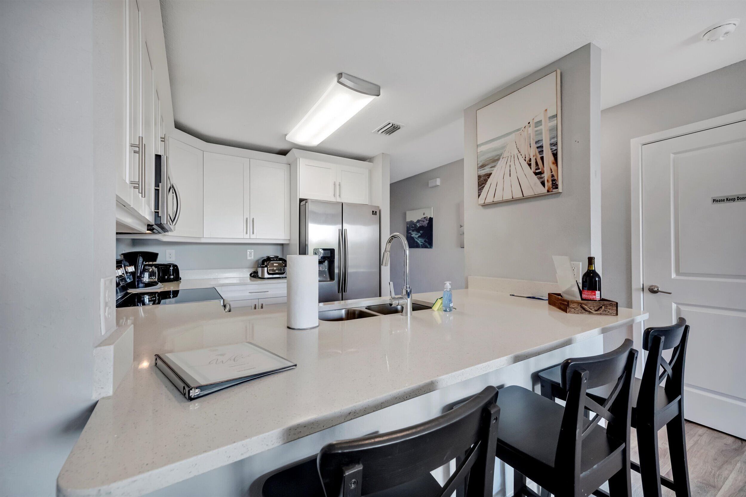 Une cuisine moderne avec des meubles blancs, un réfrigérateur en acier inoxydable, un coin bar et un espace repas avec des chaises noires.
