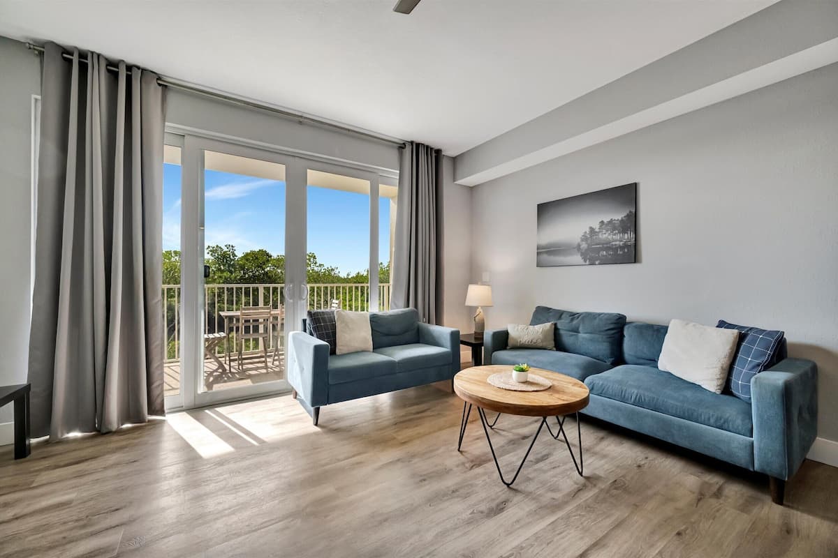 A modern living room with a blue sofa, a round wooden coffee table, and a balcony with a view of greenery.
