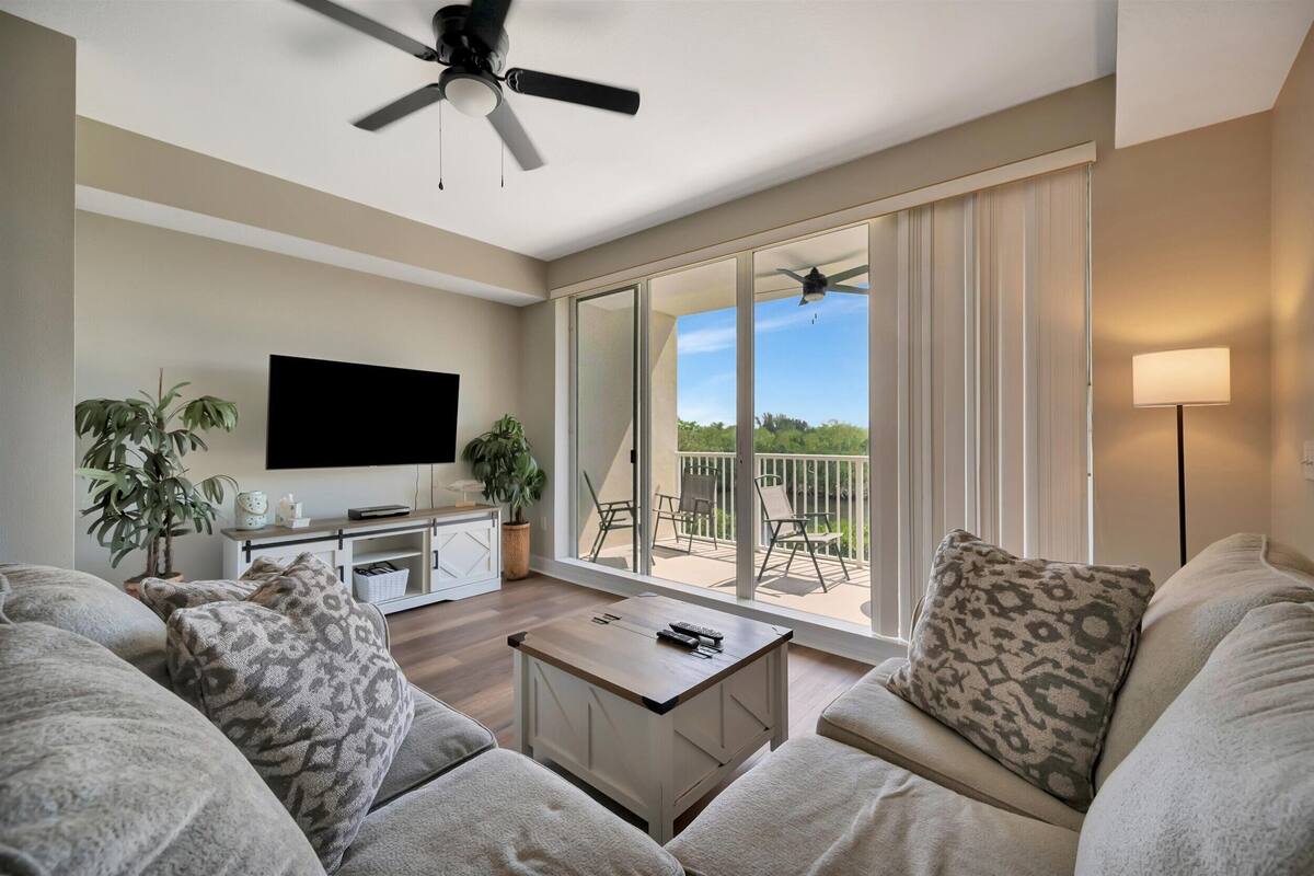 A living room with a flat-screen TV, a ceiling fan, a coffee table, and a balcony with outdoor furniture.