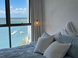 A bedroom with a large window overlooking a body of water, a bed with patterned bedding, and a hanging light fixture. - Waterfront Gold Coast Luxury apartment on the Lagoon (Labrador)