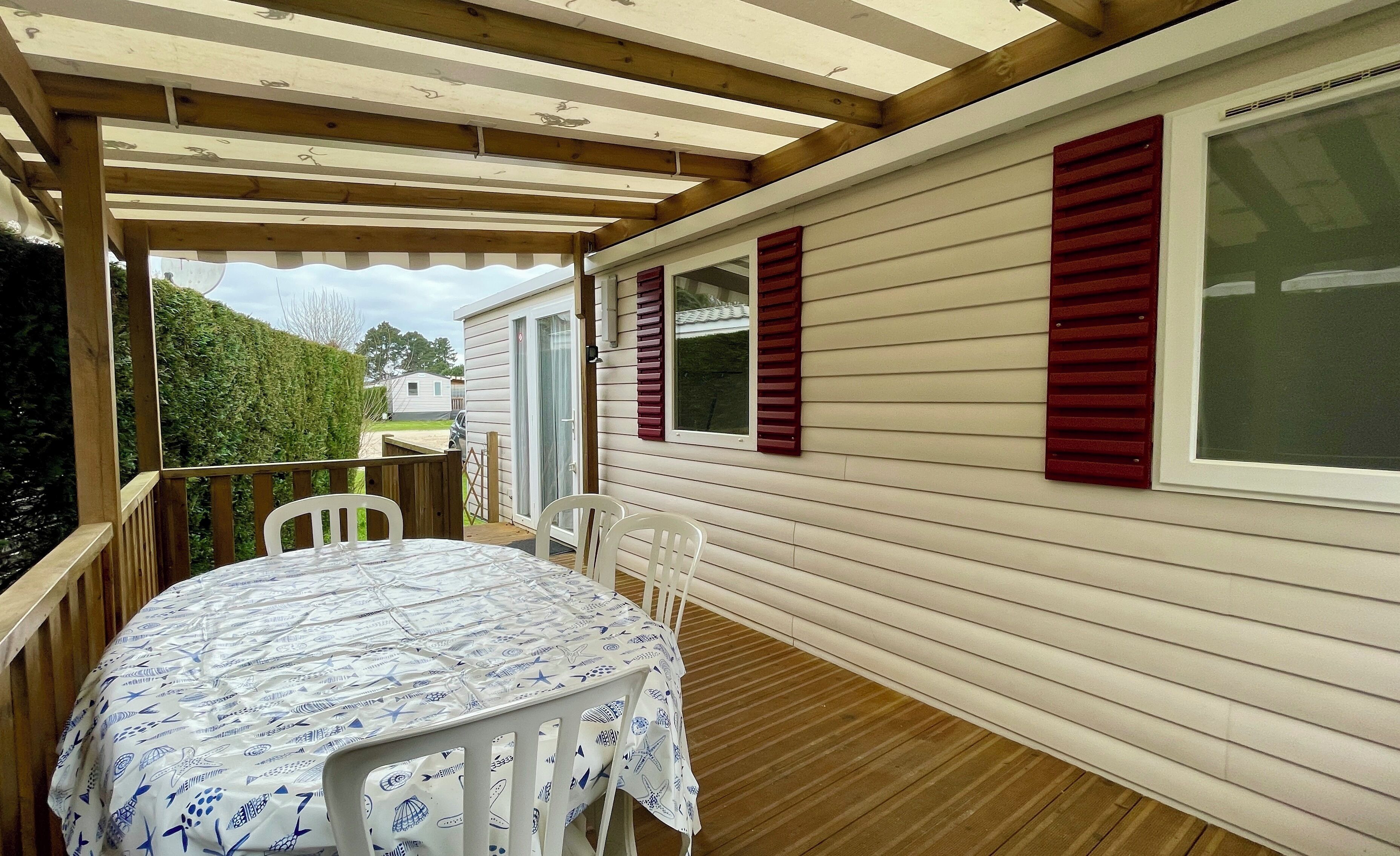 Une terrasse extérieure couverte, dotée d’une table et de chaises, de rampes en bois et offrant une vue sur une maison aux volets rouges.