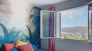 Ein Schlafzimmer mit einem Bett, farbenfrohen Kissen, einem Fenster mit Blick auf ein Gewässer und Gebäude sowie einem Wandbild mit Palmen.