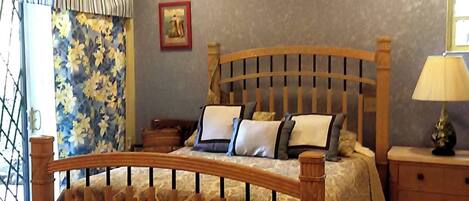 A bedroom with a wooden bed, floral curtains, a bedside table with a lamp, and a wall-mounted air conditioner.