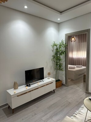 A modern living room with a flat-screen TV on a white console, a large plant, and a bed with a canopy. - Cozy apartment very close to the beach (Hammamet Sud)