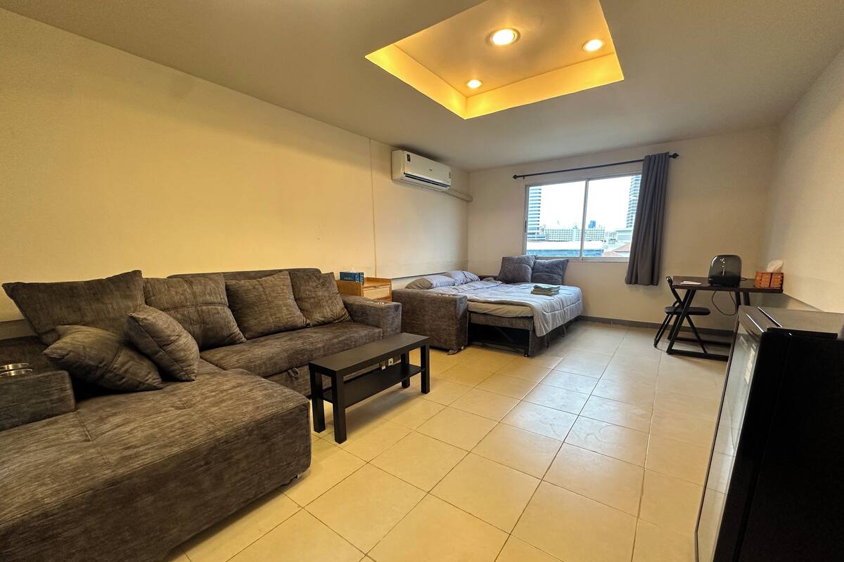A living room with a sofa, a small table, a bed, a desk, and a window with curtains.