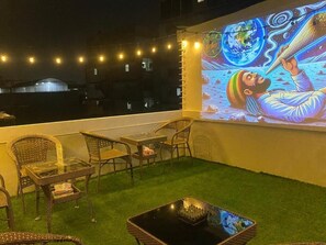 An outdoor seating area with wicker furniture, a glass table, and a projection screen displaying a space-themed image. - Holy Hostel Sathorn (Bangkok)