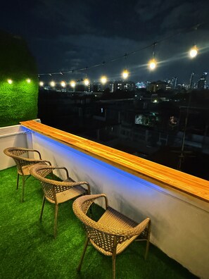 A rooftop bar with wicker chairs, a wooden counter, and a view of a cityscape at night.