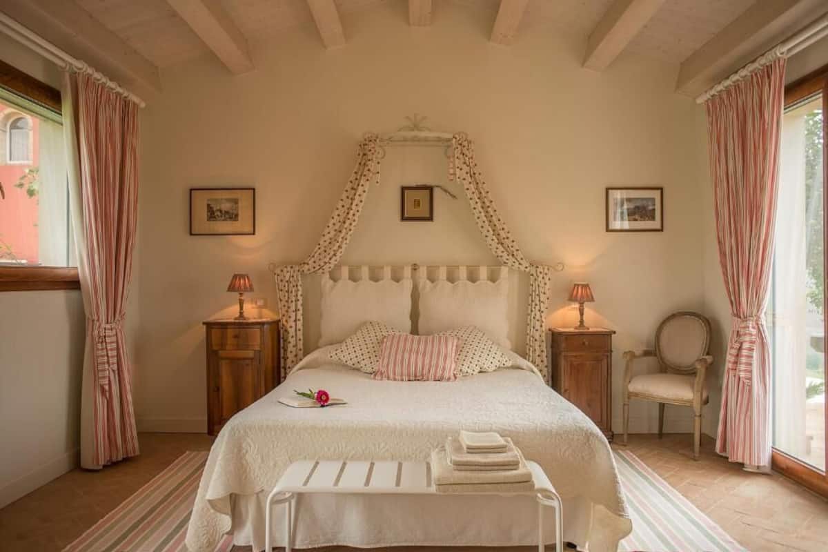A bedroom with a canopy bed, two bedside tables with lamps, a chair, and a window with curtains.