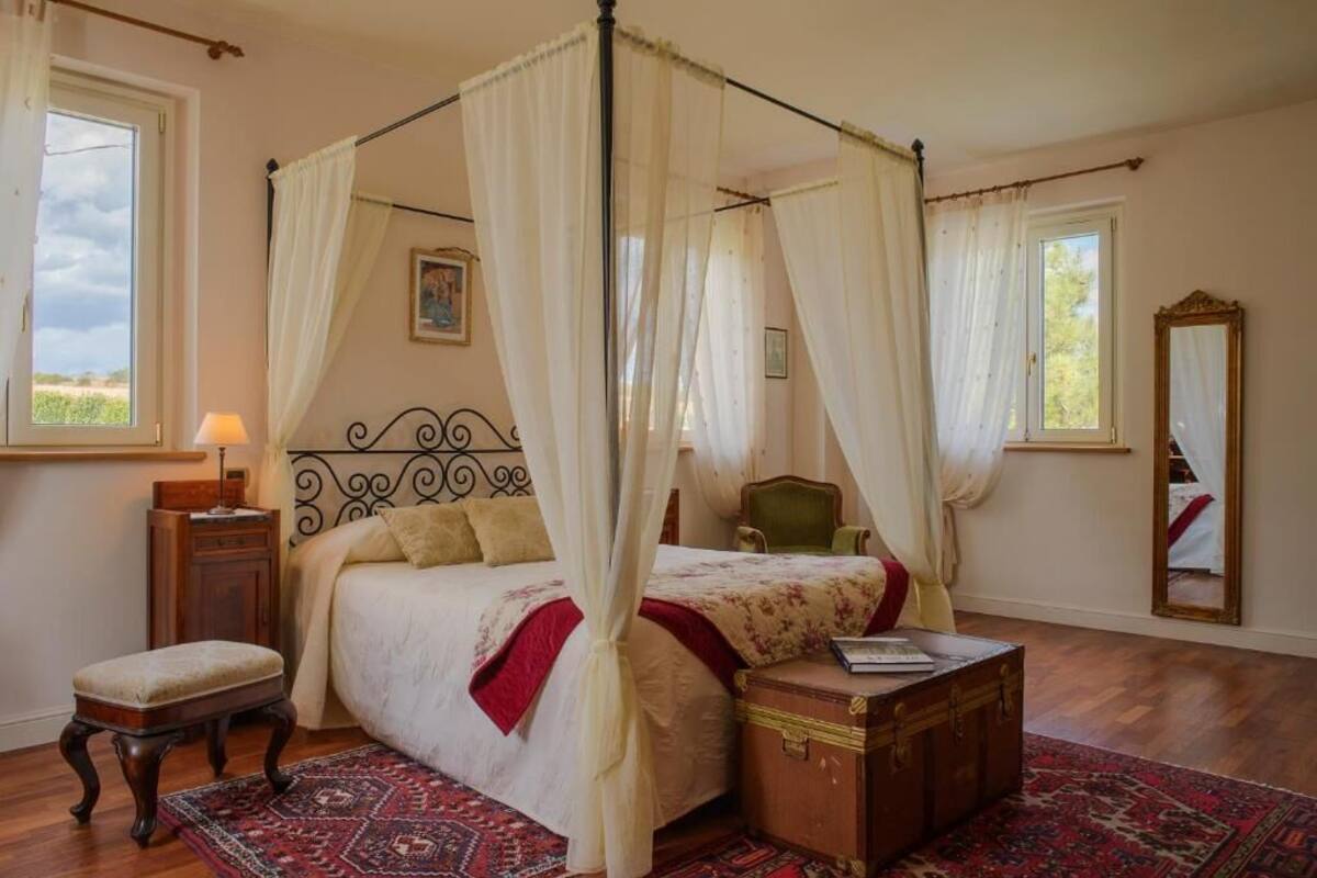 A bedroom with a four-poster bed, a nightstand, a chair, a wooden chest, and a large mirror.