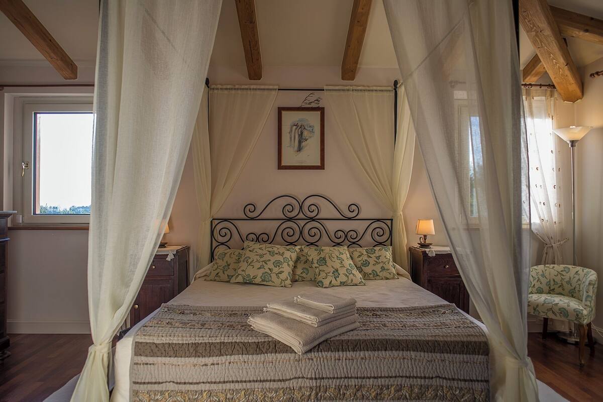 A bedroom with a four-poster canopy bed, a framed artwork above the bed, and a window with curtains.
