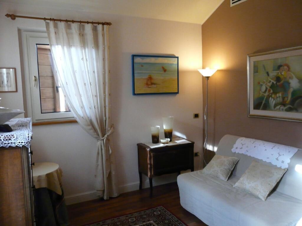 A bedroom with a bed, a nightstand with a lamp, a window with curtains, and a painting on the wall.