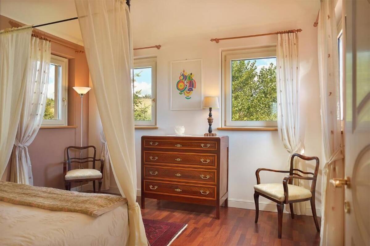 A bedroom with a bed, a dresser, two chairs, a window with curtains, and a wall-mounted lamp.