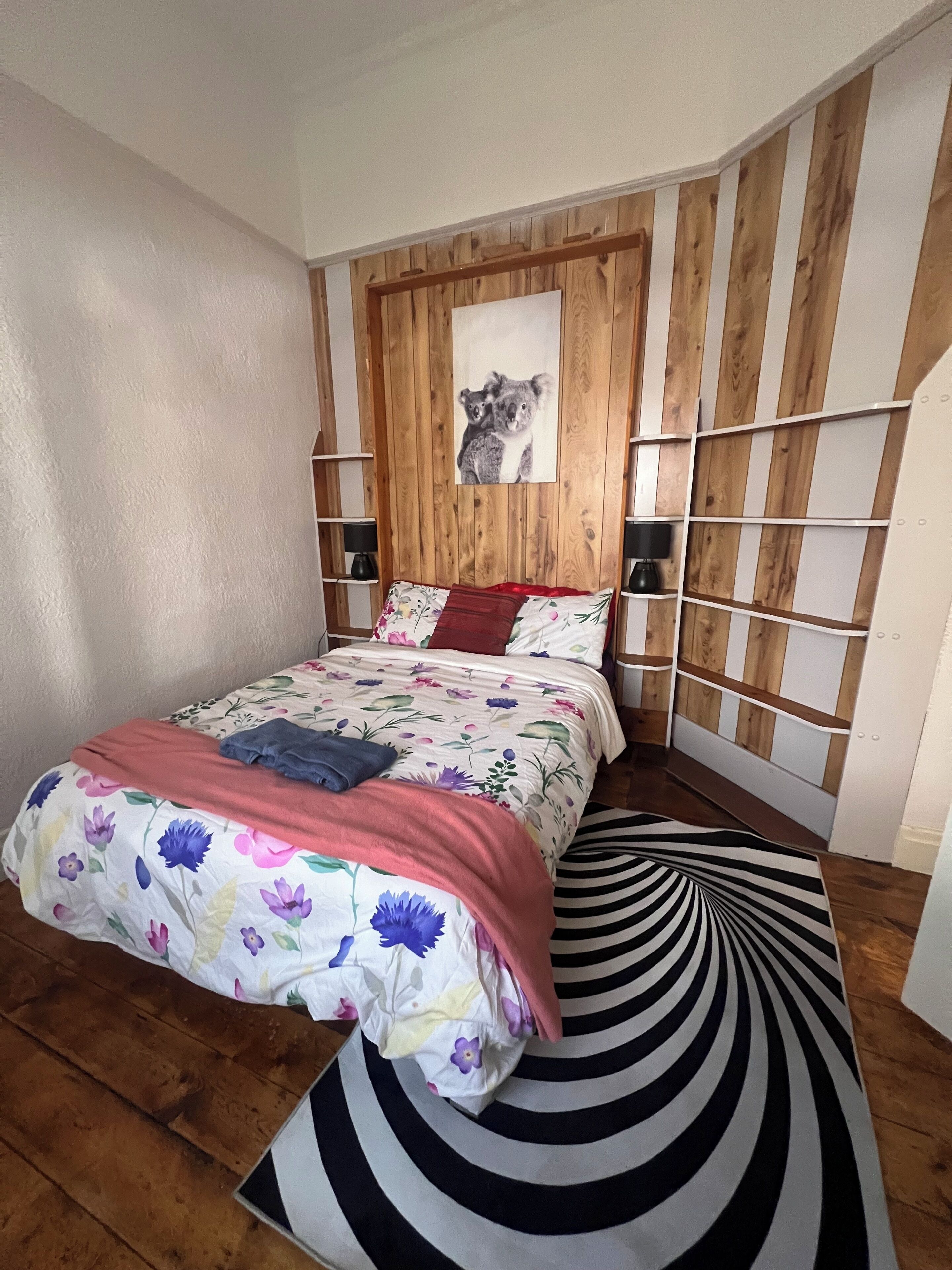 A bedroom with a wooden headboard, a floral bedspread, a black and white striped rug, and a koala-themed wall art.