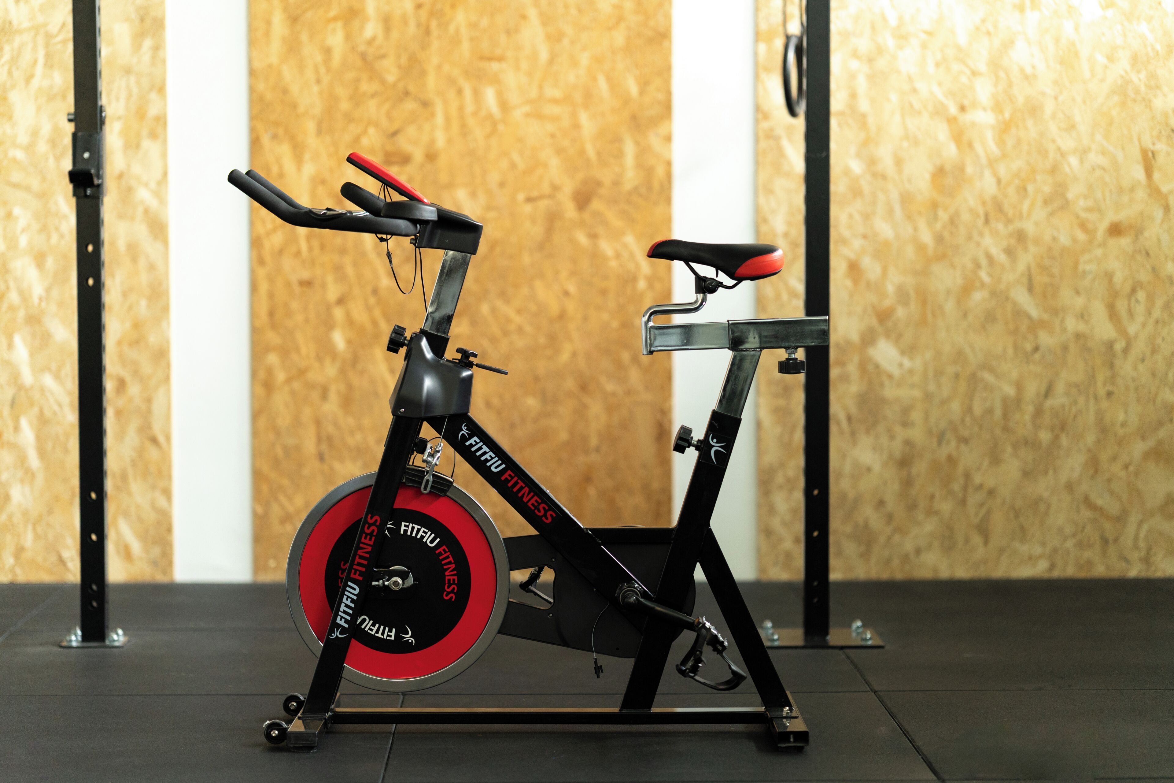 Uma bicicleta ergométrica com assento vermelho e estrutura preta, com a marca 'FITFU FITNESS', em um quarto com parede de madeira e suporte metálico.
