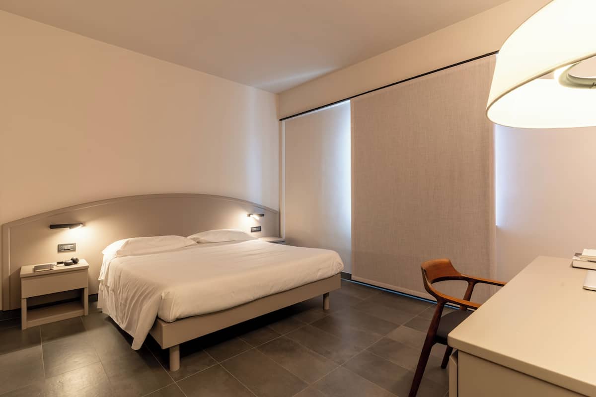A modern hotel room with a bed, bedside table, lamp, chair, and a desk.
