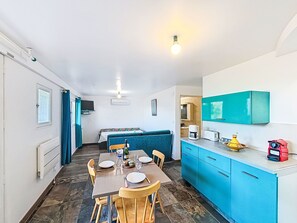 Ein gemütlicher Raum mit Esstisch, Stühlen, einer Küchenzeile, einem Bett mit blauer Kopfteil und einer Deckenleuchte.