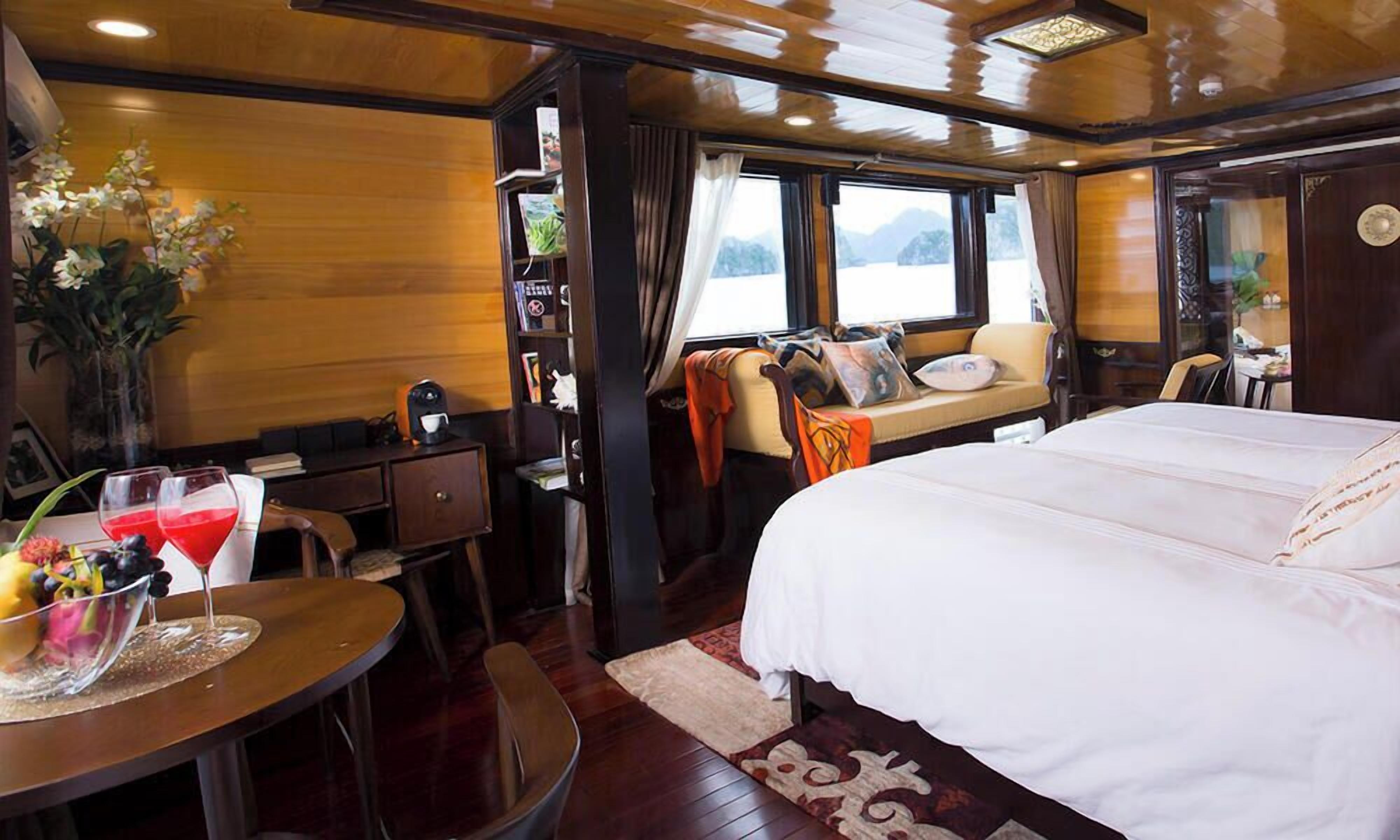 A room with a bed, a desk, a chair, a table with fruit and drinks, and a window with curtains.