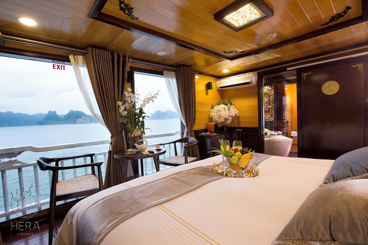 Habitación de crucero con cama, una mesa con fruta, ventana con cortinas y vista al mar.