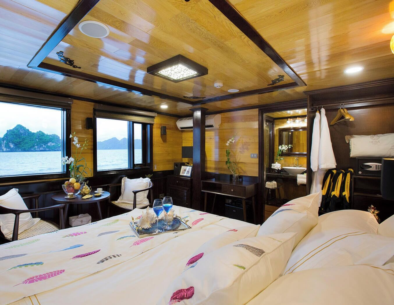 A cabin with a bed, a table with a tray of fruit, a window with a view of the sea, and a mirror.
