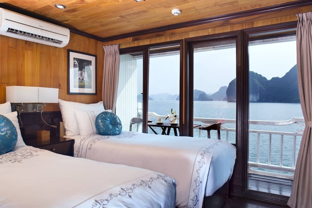 Habitación de hotel con dos camas, techo de madera, aire acondicionado, un cuadro enmarcado, una mesita con sillas y vistas al mar y a las montañas.