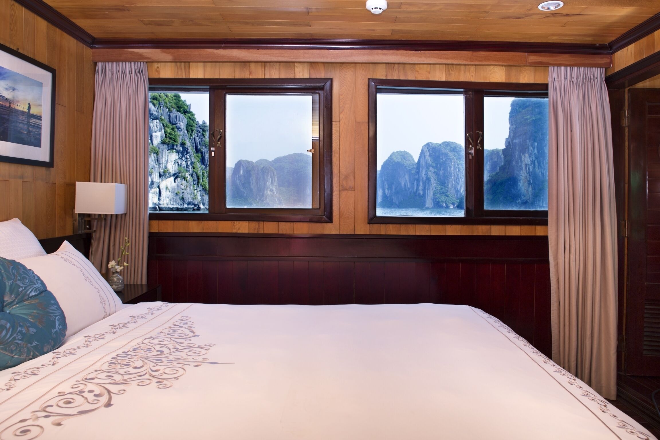 A hotel room with a large bed, wooden paneling, two windows with curtains, and a view of cliffs.