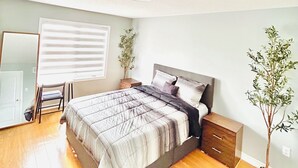 A bedroom with a bed, a nightstand, a dresser, a window with blinds, and a potted plant.