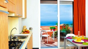 A kitchen with a sink, stove, and cabinets, a dining area with a table and chairs, and a sliding glass door leading to an outdoor patio with a view of the ocean. - Apartment in Cogoleto with pool (Cogoleto)
