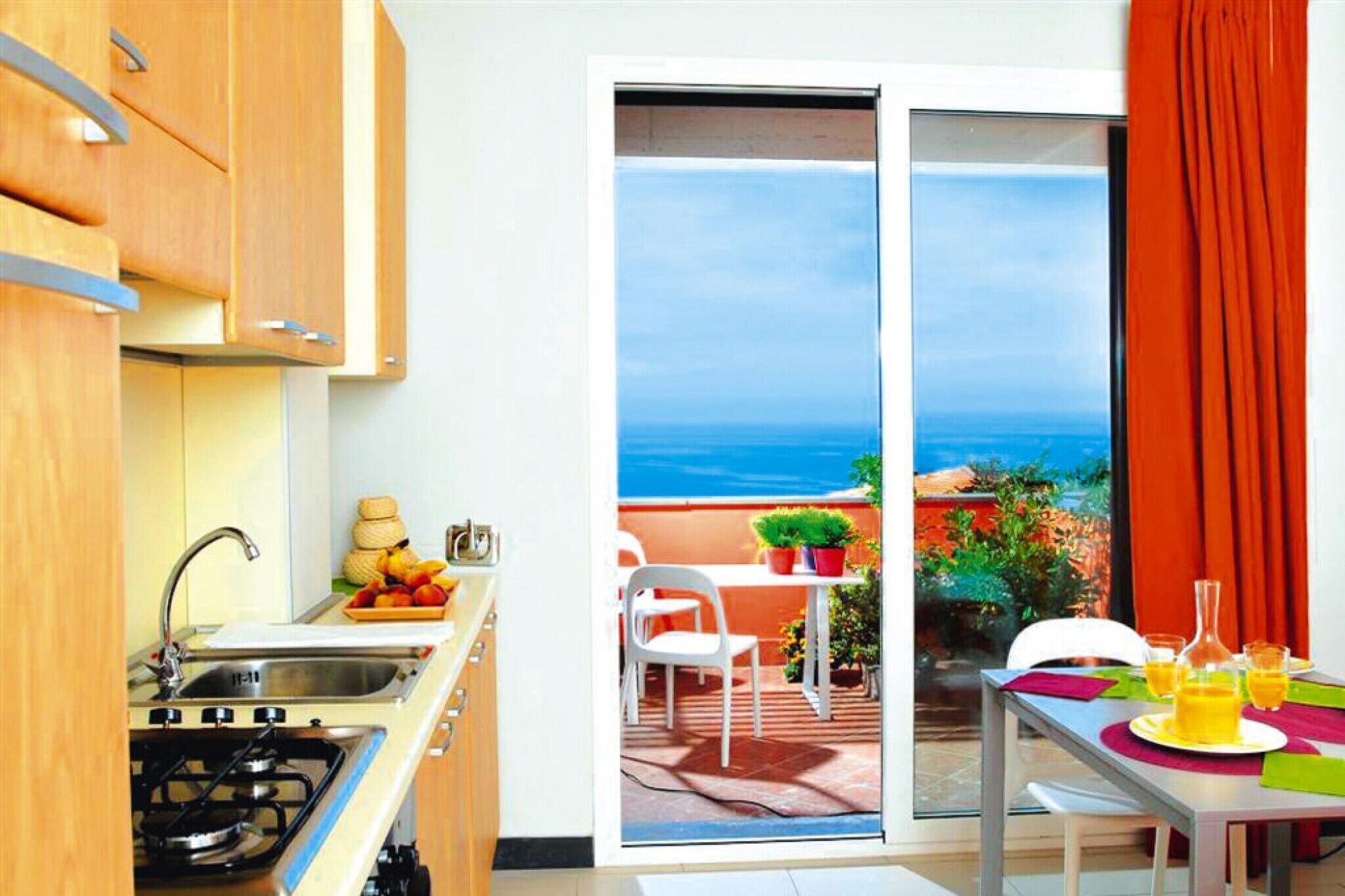 Cucina con lavello, fornelli e pensili, zona pranzo con tavolo e sedie, e una porta scorrevole in vetro che dà su un patio esterno con vista sull'oceano.