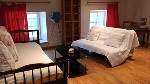 A room with a bed, a white sofa, a small table, a floor lamp, and a window with red curtains. - Family apartment with kitchen (REMONCOURT)