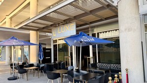 Mobiliario exterior con mesas y sillas, un cartel que dice 'LUXE LOUNGE' y un paraguas con el estampado 'CARIBBEAN TWIST'.
