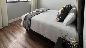 Un dormitorio moderno con una cama grande, una mesita de noche con lámpara y vistas a edificios a través de la ventana.
