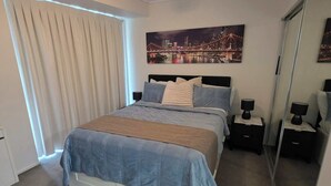 Un dormitorio con cama, ventilador de techo, mesita de noche, espejo y un cuadro de una vista urbana en la pared.