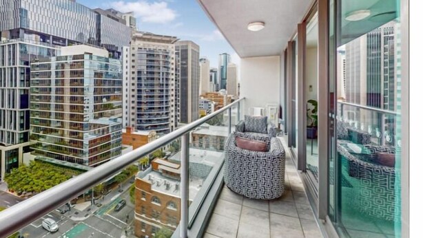 Un balcón con barandilla de vidrio, un reposapiés estampado y vistas de modernos edificios altos.