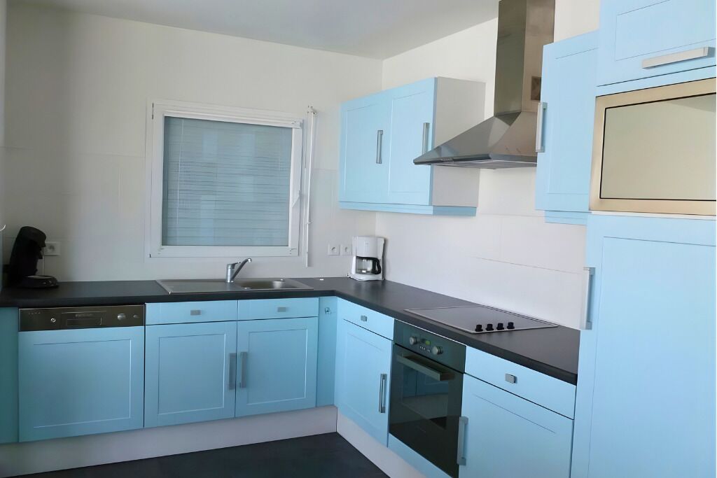 Une cuisine avec des meubles de couleur bleu clair, un plan de travail noir, une fenêtre avec des stores, une machine à café et une cuisinière.