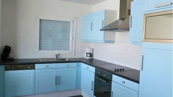 Une cuisine avec des meubles de couleur bleu clair, un plan de travail noir, une fenêtre avec des stores, une machine à café et une cuisinière.