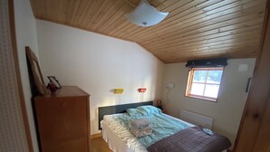 Un dormitorio con techo de madera, una cama con colcha, una mesita de noche, un aparador y una ventana con persianas.