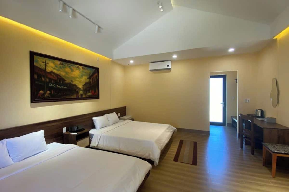 Quarto de hotel com duas camas, uma escrivaninha, uma cadeira, um quadro e uma porta que dá para outro espaço.