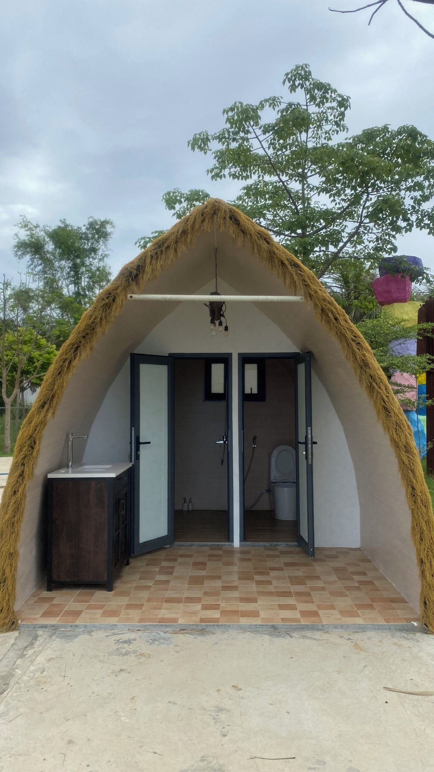 A thatched-roof structure with a sink and shower, surrounded by trees.