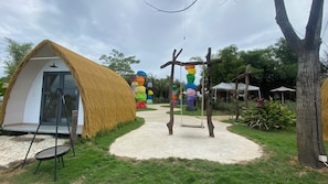 Una estructura de techo de paja con puerta de cristal, un columpio y una escultura colorida en un jardín.