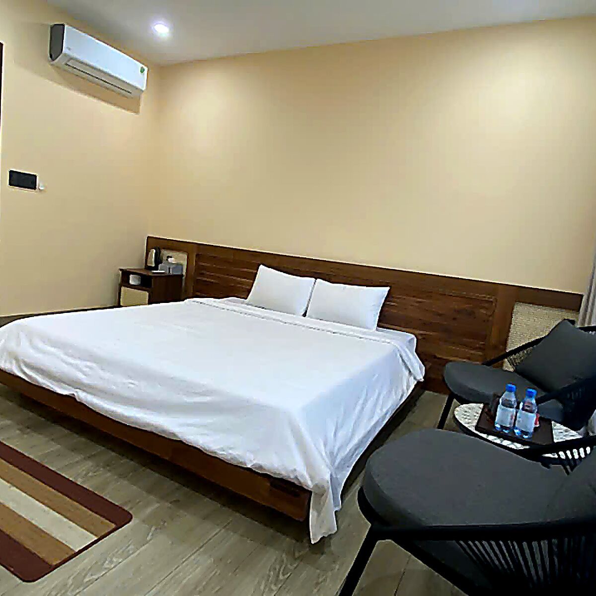 A hotel room with a bed, bedside table, chair, and a wall-mounted air conditioner.