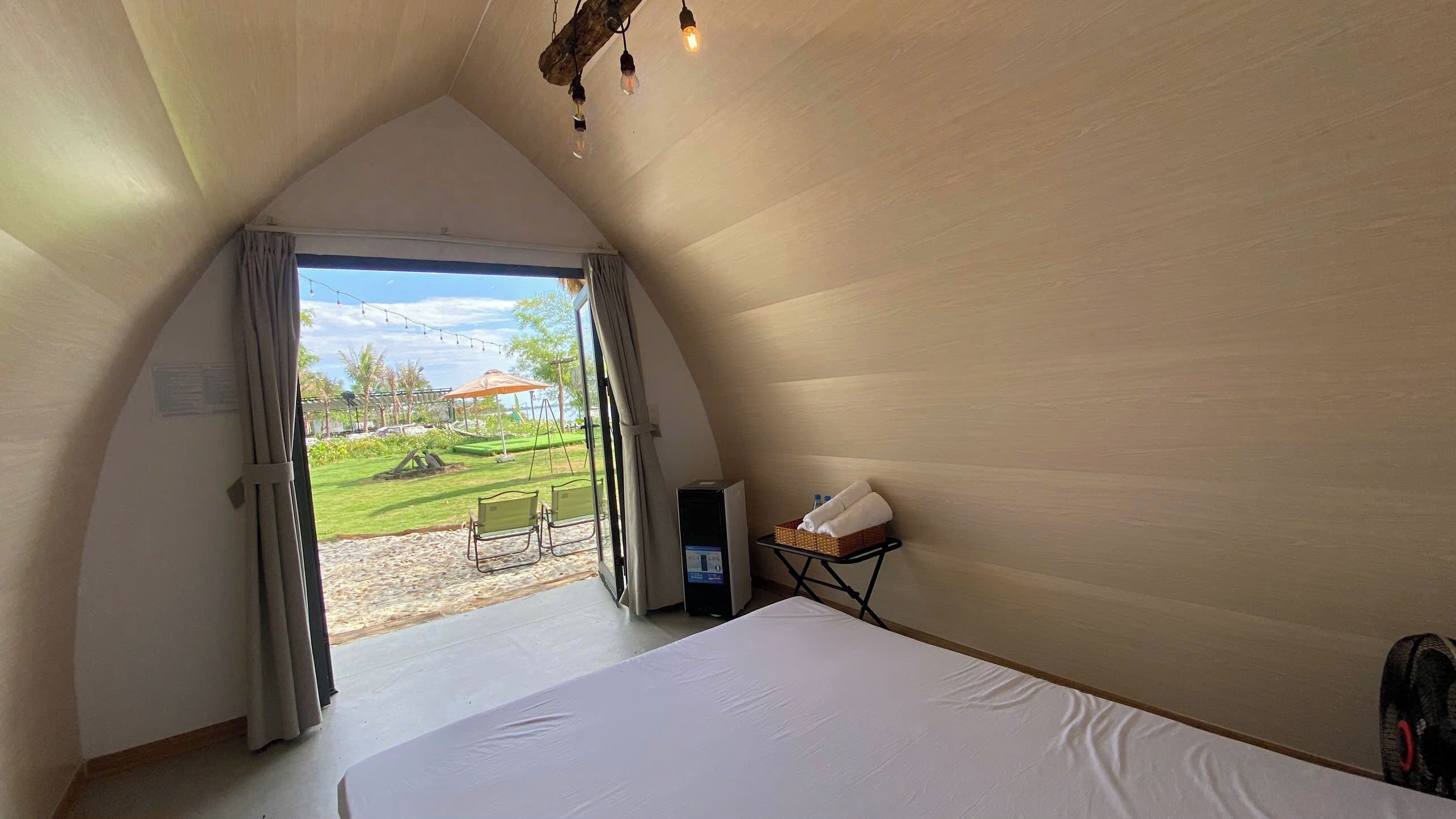 A room with a large bed, a table with a telephone, and a view of a grassy area outside.