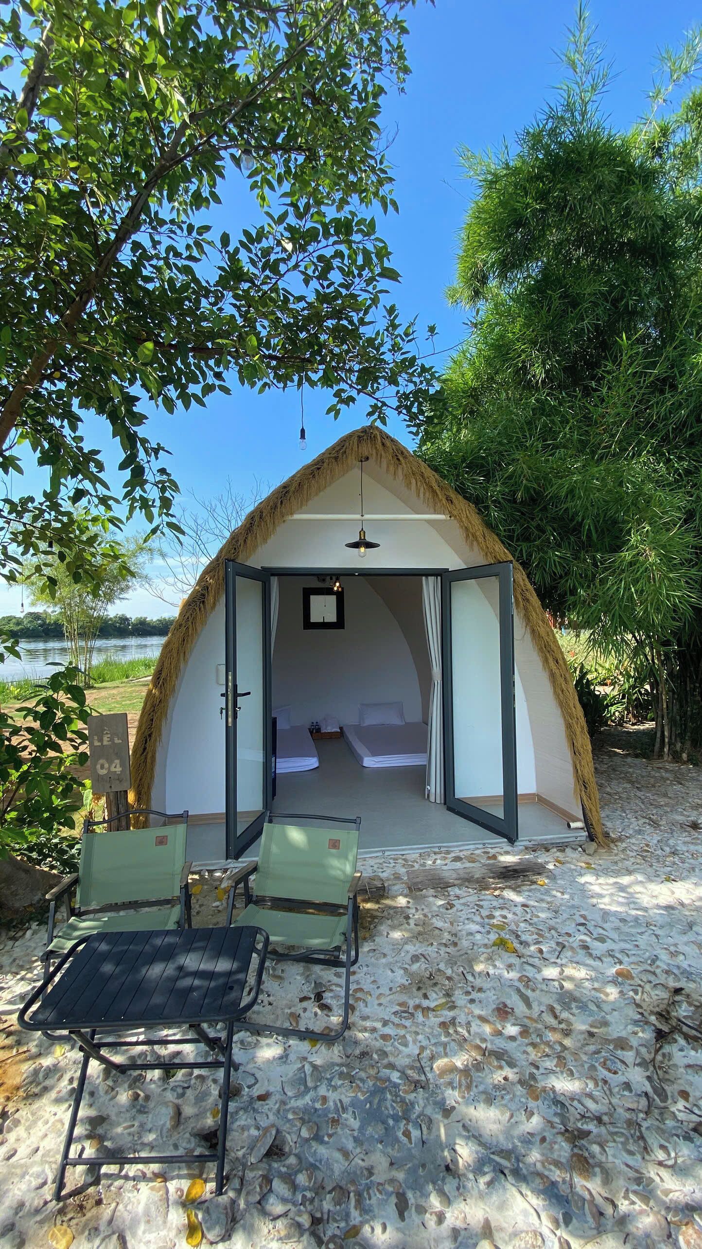 Tienda con techo de paja, con dos camas, una mesita y una silla, sobre una playa arenosa con árboles y cielo despejado.
