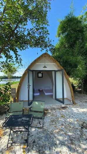 Tienda con techo de paja, con dos camas, una mesita y una silla, sobre una playa arenosa con árboles y cielo despejado.