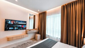 Ein modernes Hotelzimmer mit einem Wand-Fernseher, einem Holzkonsolentisch, einem großen Bett mit grauer Strukturdecke und einem Fenster mit Vorhängen.