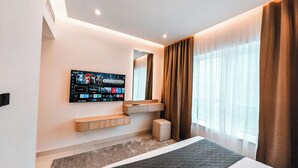 Ein modernes Hotelzimmer mit einem großen Flachbildfernseher an der Wand, einem Holzkonsolentisch, einem kleinen Hocker und einem großen Fenster mit Vorhängen.