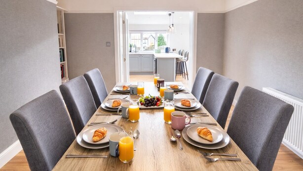 A dining area with a long table set for breakfast, chairs, and a view into the kitchen. - Stylish Skipton Retreat | Private Garden | Parking | Hiking | Yorkshire Dales (Skipton)