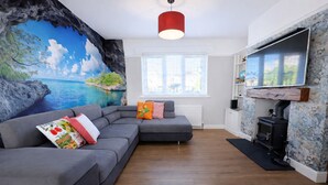 A living room with a grey sectional sofa, a fireplace, a mural of a beach scene, and a window with blinds. - Stylish Skipton Retreat | Private Garden | Parking | Hiking | Yorkshire Dales (Skipton)